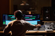 © pressmaster - Black young adult man wearing headphones working at audio mixing console in professional recording studio, adjusting sound levels on computer monitors with music producer in background