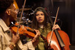 © pressmaster - Hispanic girl playing cello while looking at young Black man playing violin in music studio, both musicians focused on instruments, musical collaboration in progress