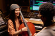 © pressmaster - Young adult Hispanic woman smiling while playing cello in music studio, interacting with young adult Black man sitting nearby, professional audio equipment visible in background
