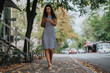 © qunica.com - A young woman in a striped dress walks on a leaf-covered sidewalk, focused on her smart phone. City life and technology blend in an autumn setting with cars in the background.