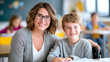 © zamuruev - Woman with glasses smiles while sitting beside young boy in classroom. Bright space filled with educational materials and engaged students in background