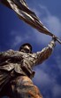 © Fathor - A monument of an Asian soldier holding up the flag, against a blue sky background, shot from a low angle with a wide-angle lens, in a sculptural style with bronze material, displaying a strong contras