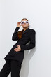 © SHOTPRIME STUDIO - Fashion woman dressed in sleek black outfit poses confidently with white sunglasses against minimalist white background, showcasing modern style and sophistication.