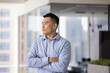 © fizkes - Serous Chinese professional man dressed in shirt standing alone in contemporary office with arms-crossed, looks into distance, thinks on business perspectives, search issue solution, making decision