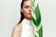 © VICHIZH - Young woman holding a large green leaf, showcasing a natural and serene beauty in a minimalistic white outfit against a soft background.