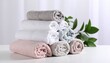 © Muji Creatives - Stack of rolled towels in white, light pink, and gray on white surface with green leaves and blurred potted plant background