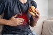 © panadda design - Asian Man Suffering from Stomach Pain and Discomfort After Eating Junk Food, Holding a Burger in Hand Experiencing Heartburn and GERD Symptoms