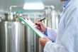 © Siyam - Scientist in lab coat writing on clipboard in laboratory with large tanks in the background