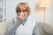 © Serhii - Senior sick frozen woman seated on sofa in living room covered with warm plaid sneezing holding paper napkin blow out runny nose feels unhealthy