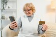 © Serhii - Portrait of happy older woman holding smartphone and credit card, engaged in online shopping activity, make digital transaction, send money through mobile application. Electronic shopping, modern tech