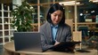 © Yuliia - Friendly Asian Korean business woman wearing headset talking laptop video call female office businesswoman Chinese Japanese ethnic girl executive writing notes information conference operator manager