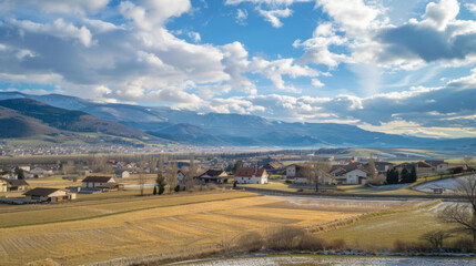scenic countryside view of a village nestled near mountains, with golden fields under a partly cloud