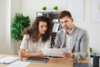 © Studio Romantic - Two young confident smiling business people and company employees in formal clothes working in office at the desk looking through financial documents on workplace discussing work project.