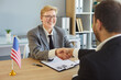 © Studio Romantic - Shaking hands across desk, people sitting greeting in office near American flag, woman smiling, happiness and hr agreement, successful meeting partnership, positive professional business interaction