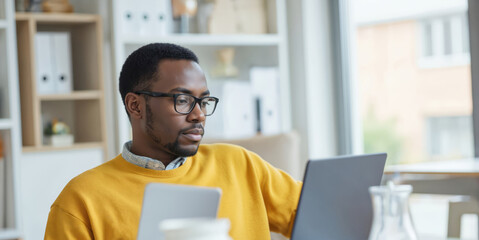 African American businessman in yellow sweater uses laptop, tablet in office. Wears glasses, earphones, appearing focused. Workspace includes desk, shelves. Represents modern tech, business strategy,