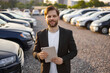 © sofiko14 - A smiling car salesman in a suit holding a tablet in a car dealership, with rows of cars in the background.