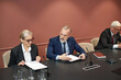 © DragonImages - Middle aged Caucasian woman and middle aged Caucasian man discussing documents at conference table with microphones, while senior Caucasian man reading papers during political meeting
