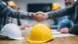 © CStock - Two professionals shake hands over construction plans, with hard hats on the table, symbolizing collaboration.