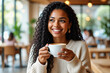 © f_bossa - A smiling young woman with curly hair holds a coffee cup while gazing thoughtfully out of a cafe window.