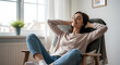 © peno - Young relaxed smiling pretty woman relaxing sitting on chair at home. Happy positive beautiful lady feeling joy enjoying wellbeing and lounge chilling near window in modern cozy apartment interior