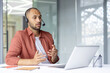 © Liubomir - A man in a headset is looking at a laptop and gesturing with his hands during a video call in a modern office setting.