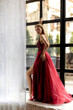 © Svetlana - Elegant woman in red gown poses gracefully by large windows in a stylish urban setting during the golden hour