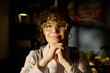 © pressmaster - Portrait of middle aged Caucasian woman with curly hair and glasses smiling gently while resting chin on hands, looking directly at camera in softly lit indoor setting