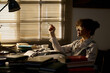 © pressmaster - Caucasian young adult woman sitting at desk holding cigarette surrounded by stacks of books and papers, wearing glasses, gazing sideways with thoughtful expression