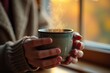 © Artem Zatsepilin - close up on hands in warm sweater holding steaming mug by window with autumn colors. warmth and comfort at cozy home. fall season, relaxation, beverage enjoyment.
