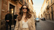 © miss irine - Elegant woman with blonde hair walks down Italian city street in autumn. Wears stylish beige suit, sunglasses, large earrings. Two men in dark coats follow. Street lined with buildings, parked cars.