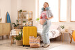 © Pixel-Shot - Young pregnant woman with headphones, travelling accessories and suitcase in bedroom