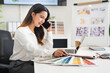 © Fahng - Concentrated young interior designer woman sitting at table with blueprint drawing ,swatches and material , discussing interior design with client  by phone, choosing materials for decorations.