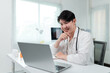 © amnaj - Asian male doctor wearing lab coat and stethoscope is making a video conference with a patient on a laptop while sitting at his desk in the hospital office