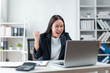 © amnaj - Young asian manager raising her fist and screaming while looking at her laptop, celebrating a successful project or achievement in her modern office workplace