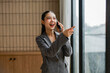 © amnaj - Smiling businesswoman wearing a gray suit is talking on her phone and pointing out the window in her office, indicating something interesting or important