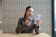 © amnaj - Smiling Asian businesswoman in suit sitting at a wooden table and having a video call with her smartphone, pointing her finger at the screen