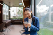 © amnaj - Young Asian businesswoman wearing blue suit using mobile phone and smiling while sitting near window in modern office on a rainy day, enjoying online communication or successful business deal