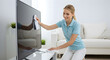 © abu - Smiling Blonde Woman Cleans Flat Screen Tv in a Bright White Living Room with Cloth Wearing Blue Polo and White Pants