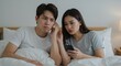 © rdkcho - Young Asian Couple in Bed Examining a Smartphone Together, Showing Signs of Relationship Trouble or Jealousy