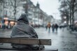 © Patrick Louvet - Homeless person wrapped in blanket sitting alone on bench during snowfall in cold winter city, highlighting urban poverty and social isolation