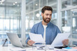 © Liubomir - A smiling businessman reviews documents at his desk in a bright, modern office setting.