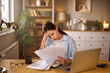 © Stockphotodirectors - A woman sits at her desk in a home office, looking stressed as she examines bills and financial papers. The lighting creates a somber mood, enhancing her feelings of concern.