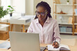 © Studio Romantic - Portrait of a concentrated serious african american business woman sitting in a shirt and glasses at the desk on her workplace and working on laptop at office looking at the monitor screen.