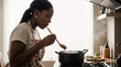 © Olga B.G. - Curvy black woman tasting sauce from wooden spoon in farmhouse kitchen
