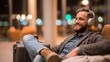 © Sergey - Bearded traveler in casual clothes, relaxing in an airport lounge with large over-ear headphones, feet up on a carry-on suitcase, ambient warm lighting