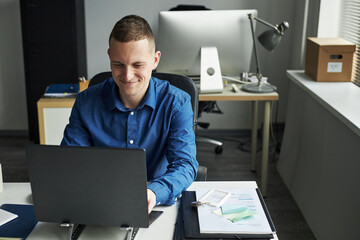 Wall Mural - Caucasian young adult man sitting at desk working on laptop in modern office, smiling while typing, documents and smartphone on table, computer monitor and office supplies in background