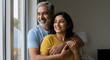 © Dual Studio - Smiling mid adult couple hugging each other and standing near window while looking outside. Happy and romantic mature man embracing hispanic wife from behind while standing at home with copy space.