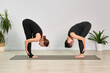 © TRAVELARIUM - Two young women in black activewear perform forward bend poses on yoga mats in minimalist studio with green plants, evoking calm and focus. Side view captures symmetry