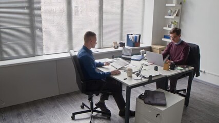 Wall Mural - PAN shot of two focused office employees typing on laptops then passing folder with documents while working at shared desk in open office space