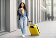 © Prostock-studio - Cheerful pretty curly middle eastern young woman in comfy casual outfit going vacation alone, walking by airport with yellow luggage, using cell phone, online check-in, copy space, full length